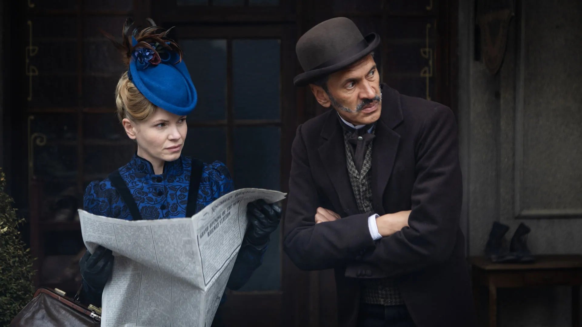 Eliza holds a newspaper while leaning forward to look at something alongside Clarence