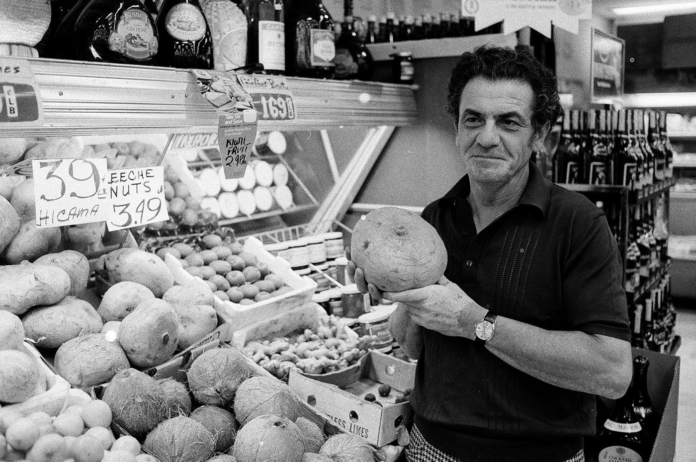 A man holds up jicama in front of a rack of fresh produce in a black and white photo