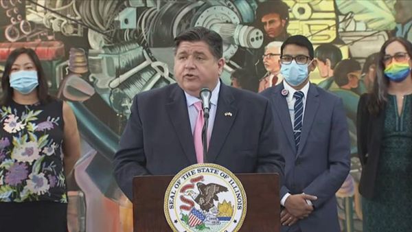 Governor J.B. Pritzker standing at a press conference, with three people wearing masks behind him