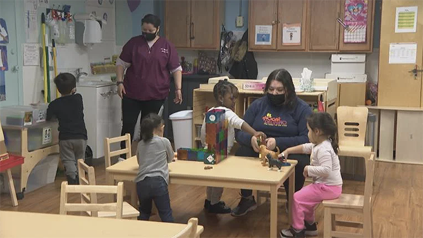 Four children and two adults in a preschool classroom, playing around a table