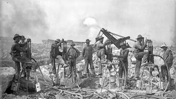 Men stand amongst broken rocks with heavy equipment in a black and white photo