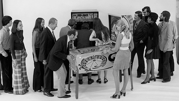 A group of people stand around two pinball machines, smiling as people play