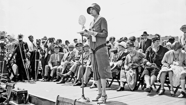 The politician Ruth Hanna McCormick stands in front of a microphone on a stage with women seated behind her and photographers pointing cameras at her in a black and white photo from the 1920s