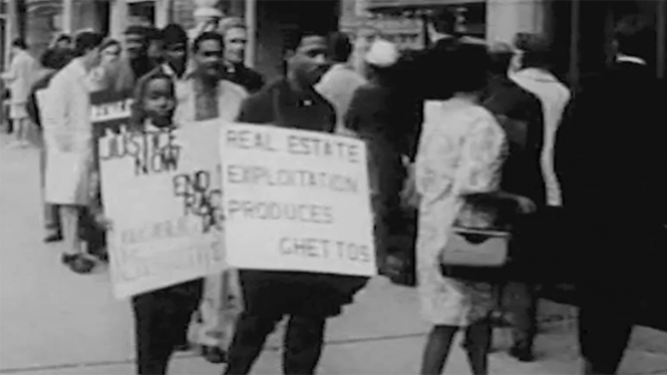 A black and white image of a man holding a sign that says, "Real Estate Exploitation Produces Ghettos"