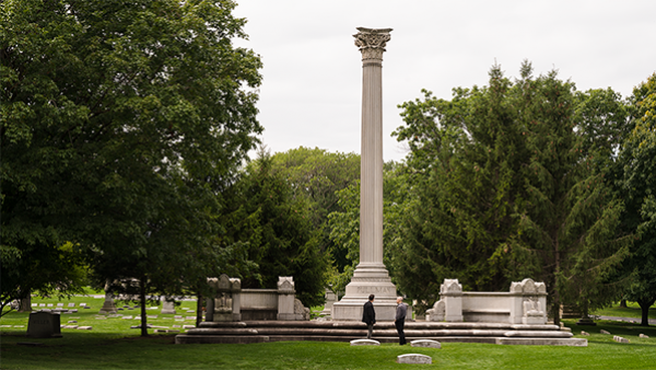 Two people stand in front of the column marking the Pullman tomb in Graceland Cemetery in Chicago
