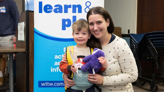 A toddler and his mother smile in front of a WTTW Kids Learn & Play banner
