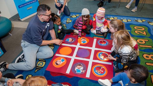 WTTW Kids Learn & Play host Cristian engages with children around a large floor game board.
