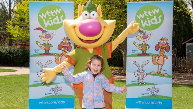 WTTW's Nature Cat and young girl smile while posing in front of WTTW Kids banners