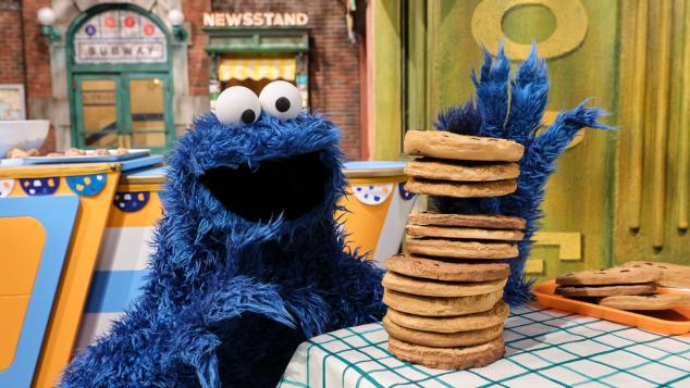 Cookie Monster sitting next to a tall stack of chocolate chip cookies on a checkered tablecloth.
