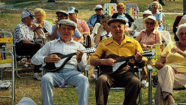Two elderly sitting in lawn chairs strumming instruments surrounded by other elderly people 