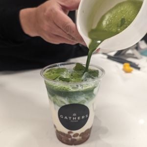 A hand pours matcha into a plastic cup on top of milk and boba