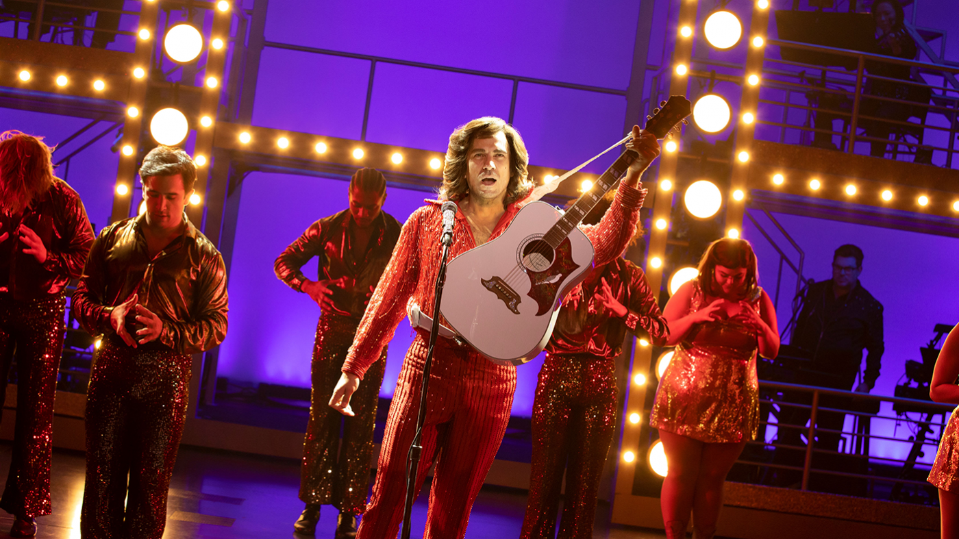 Performers on a stage, including one in sparkly clothing holding a guitar
