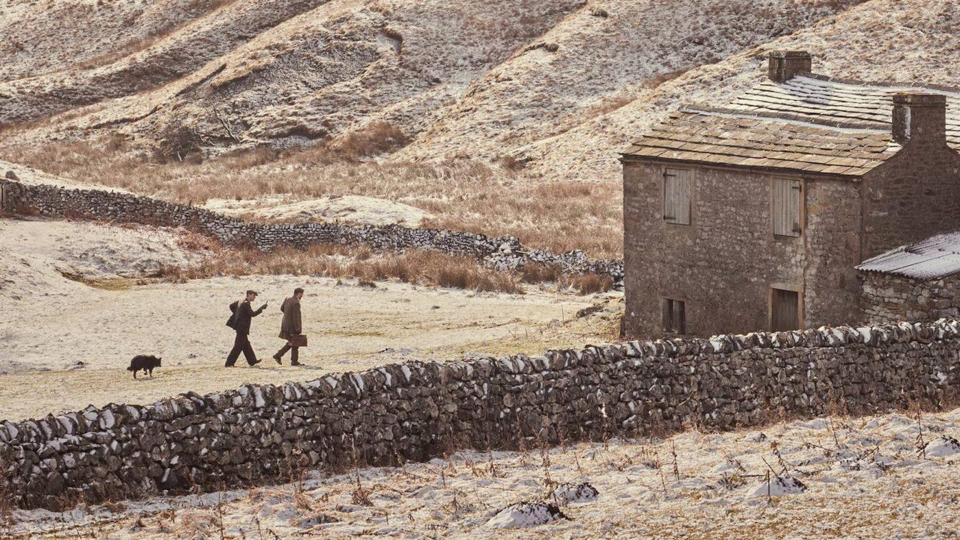 Two men and a dog walk toward a house across brown fields