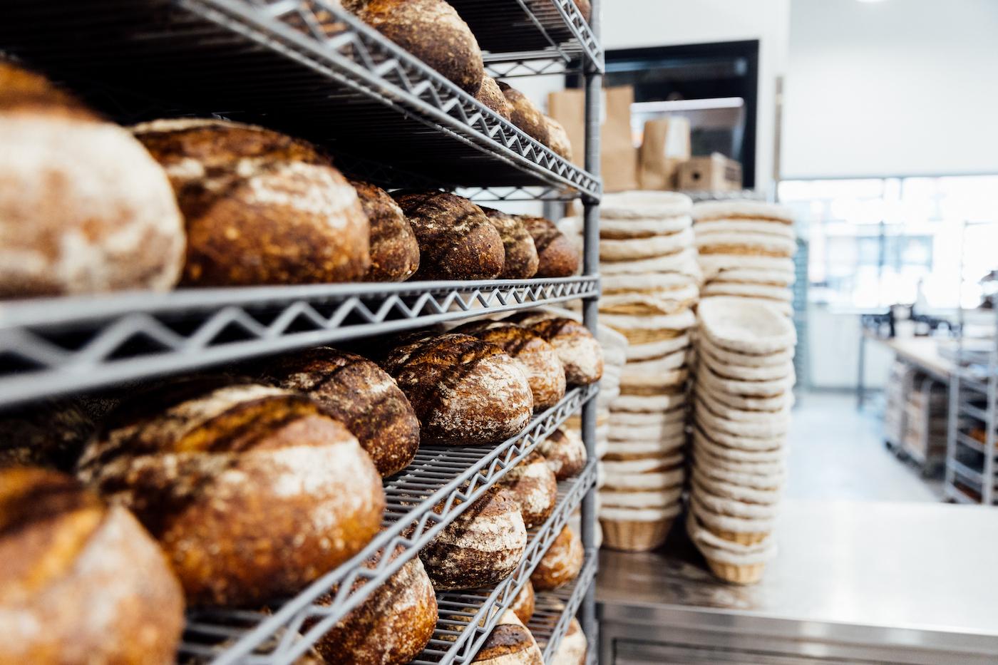 Loaves of bread cool on a wire rack in front of stacked proving baskets