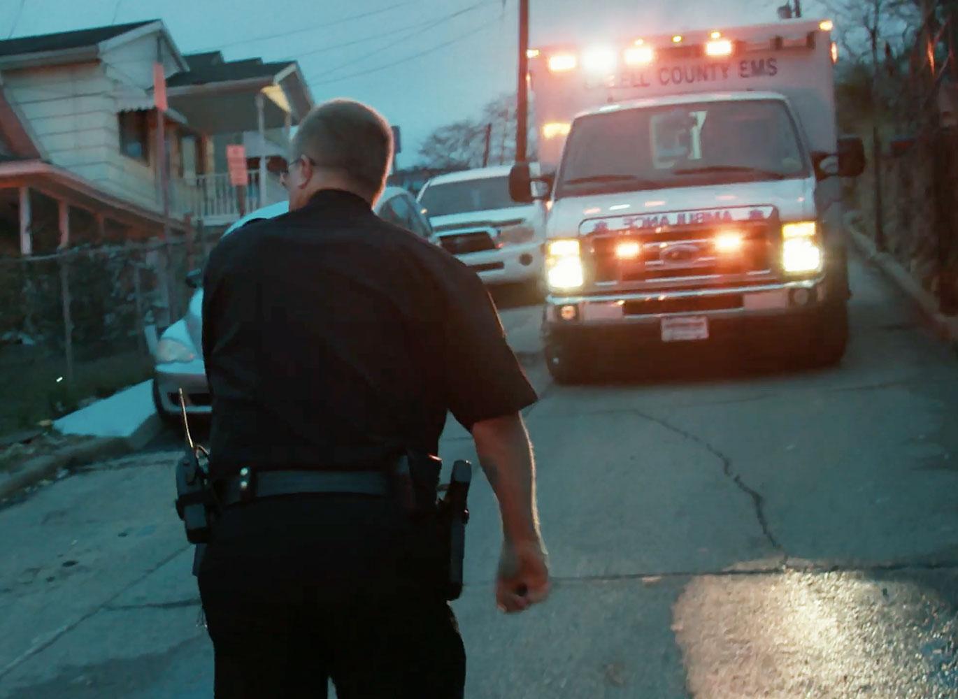 An evening scene shows a police officer walking between an police car and ambulance with flashing lights on a residential street. An evening scene shows a police officer walking between an police car and ambulance with flashing lights on a residential street.