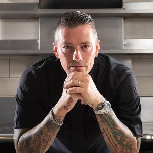 Curtis Duffy leans on his hands and stares at the camera from a stainless steel kitchen