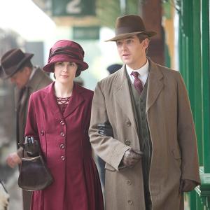 Mary and Matthew stand arm-in-arm in coats and hats at a train station
