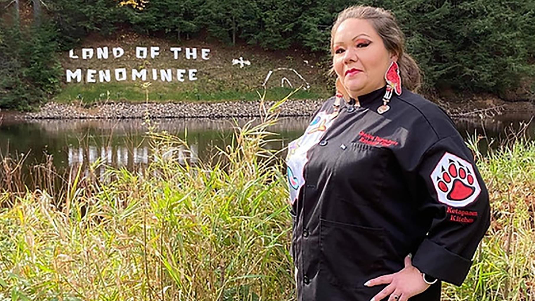 Jessica Pamonicutt standing in front of a sign on the grass reading Land of the Menominee