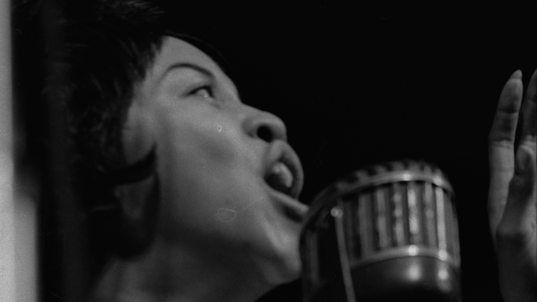 Mavis Staples singing with The Staple Singers at the second University of Chicago Folk Festival, in 1962. Photo: Chicago History Museum, ICHi-118846; Raeburn Flerlage, photographer Mavis Staples sings into a microphone with her hand up in a black and white 1962 photograph