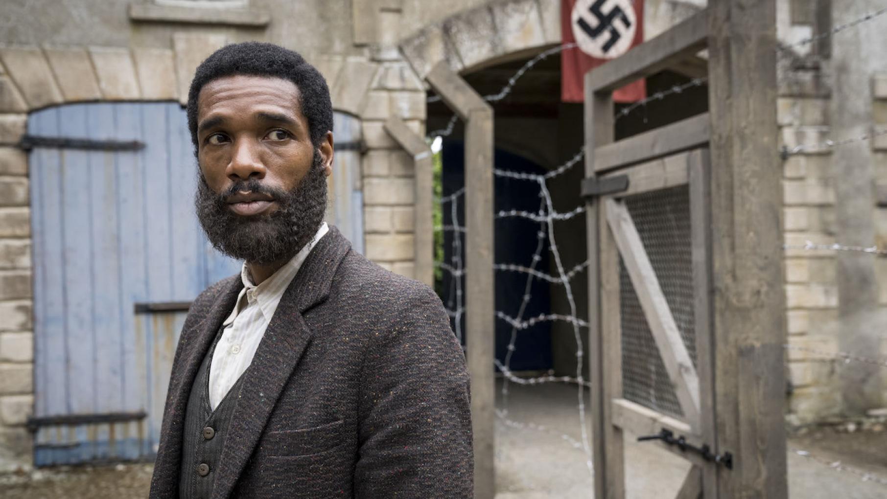 A bearded Albert stands in a prison camp with a Nazi flag in the background