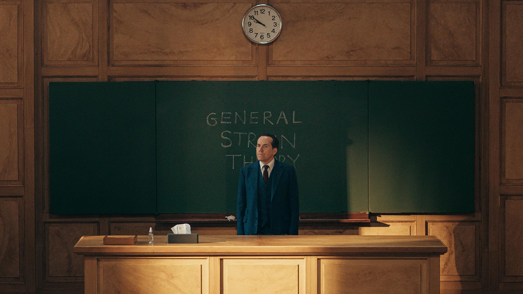 Professor T doesn't want to sit through oral exams—so he makes them extraordinarily difficult. Photo: Sofie Gheysens/Eagle Eye Drama Professor T stands behind a desk in front of a chalkboard in a lecture hall