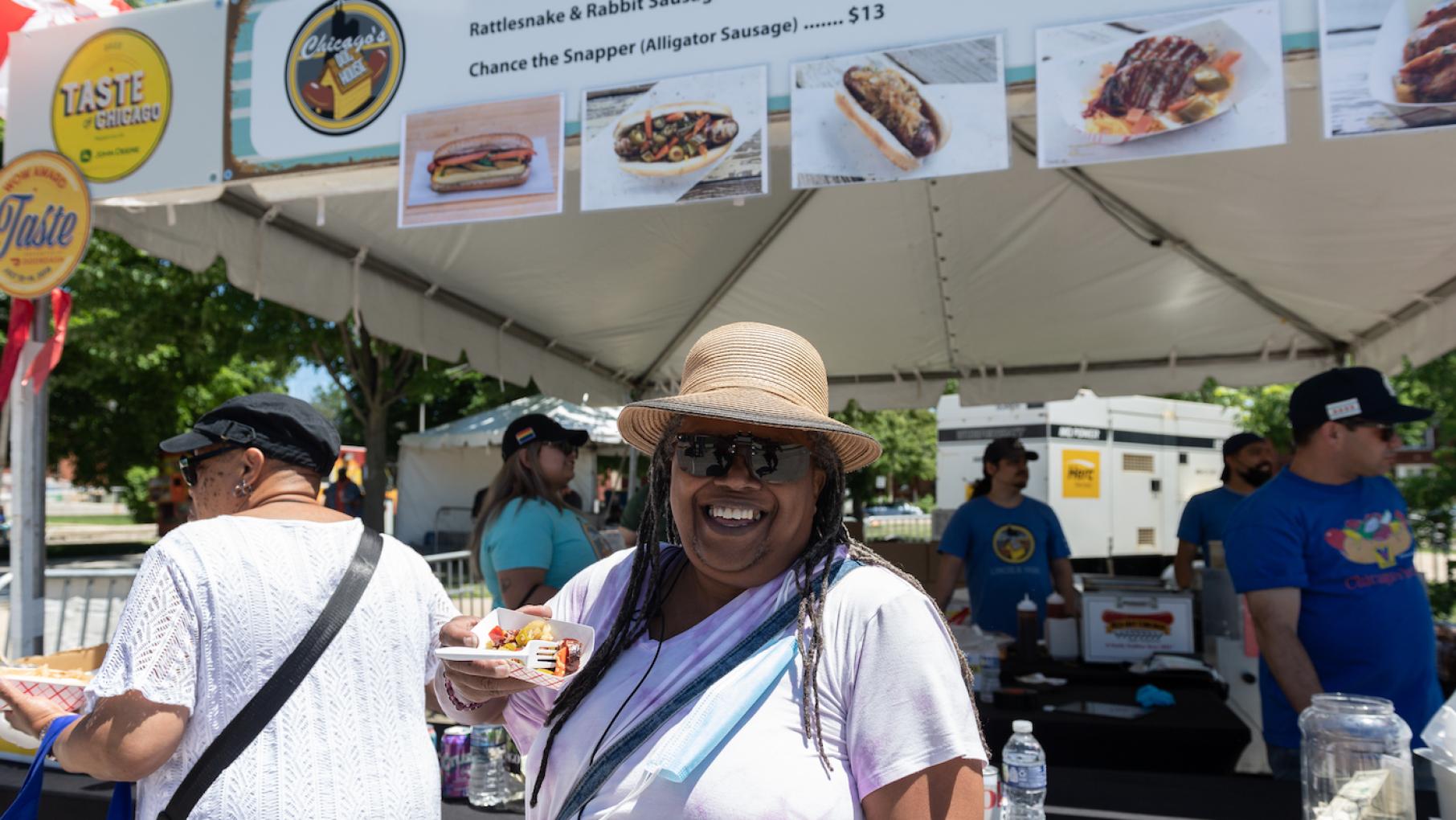 A woman smiles under a hat while holding food in front of a restaurant stand