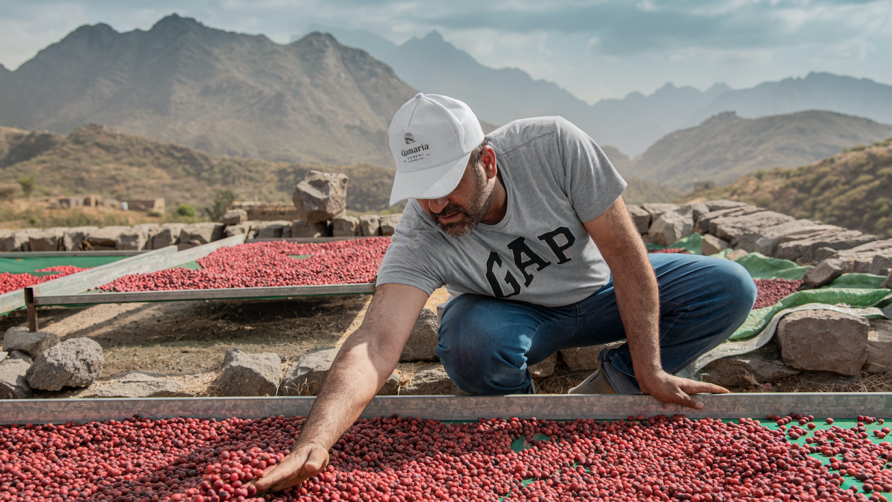 Qamaria sources its coffee from a family of four brothers in Yemen, the birthplace of coffee as a beverage. Credit: Qamaria Coffee A man squats next to a tray of coffee fruit drying in the sun in front of mountains