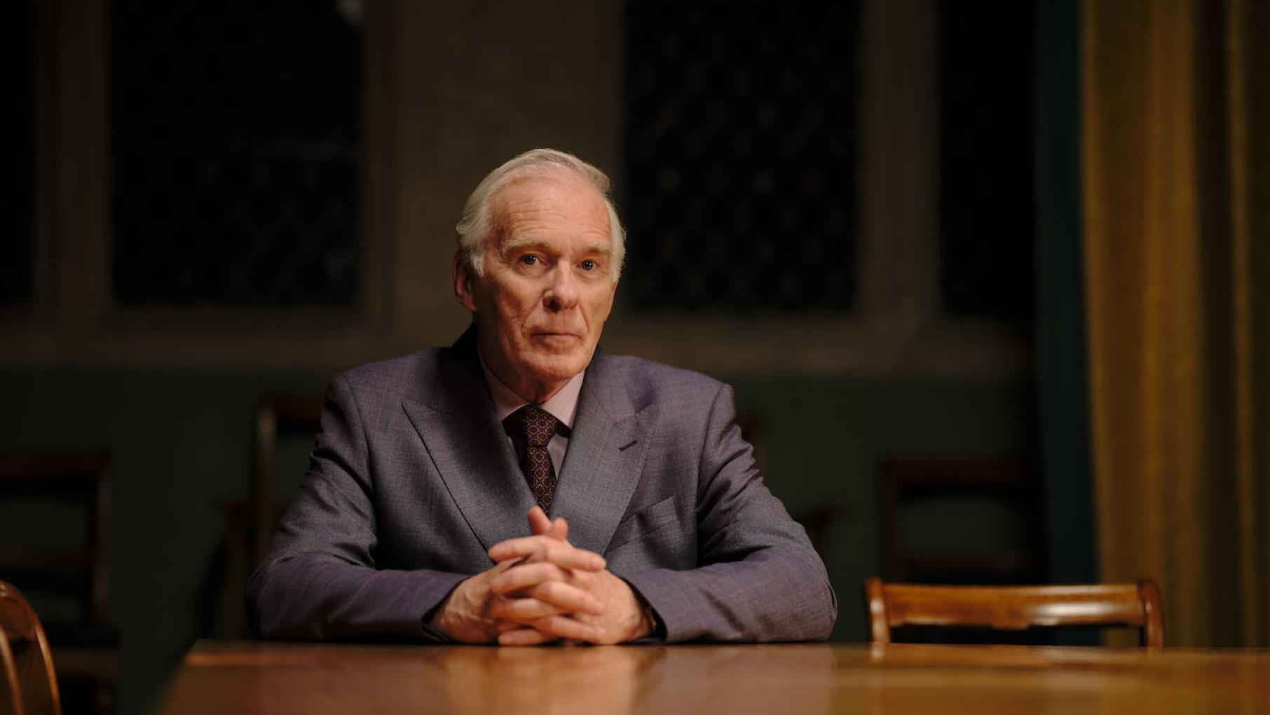 Tony Hume sits in a suit at a table, with darkness behind him