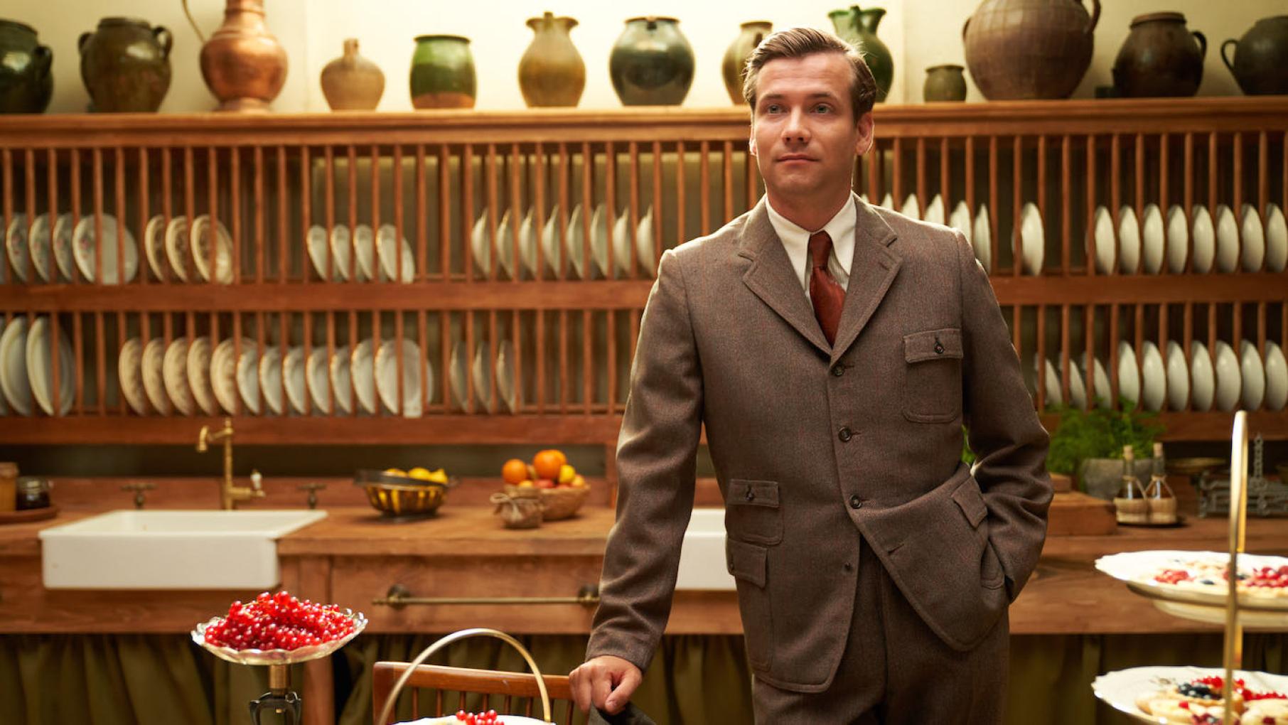 Lucian stands in a suit in front of a a wall of plates and jugs