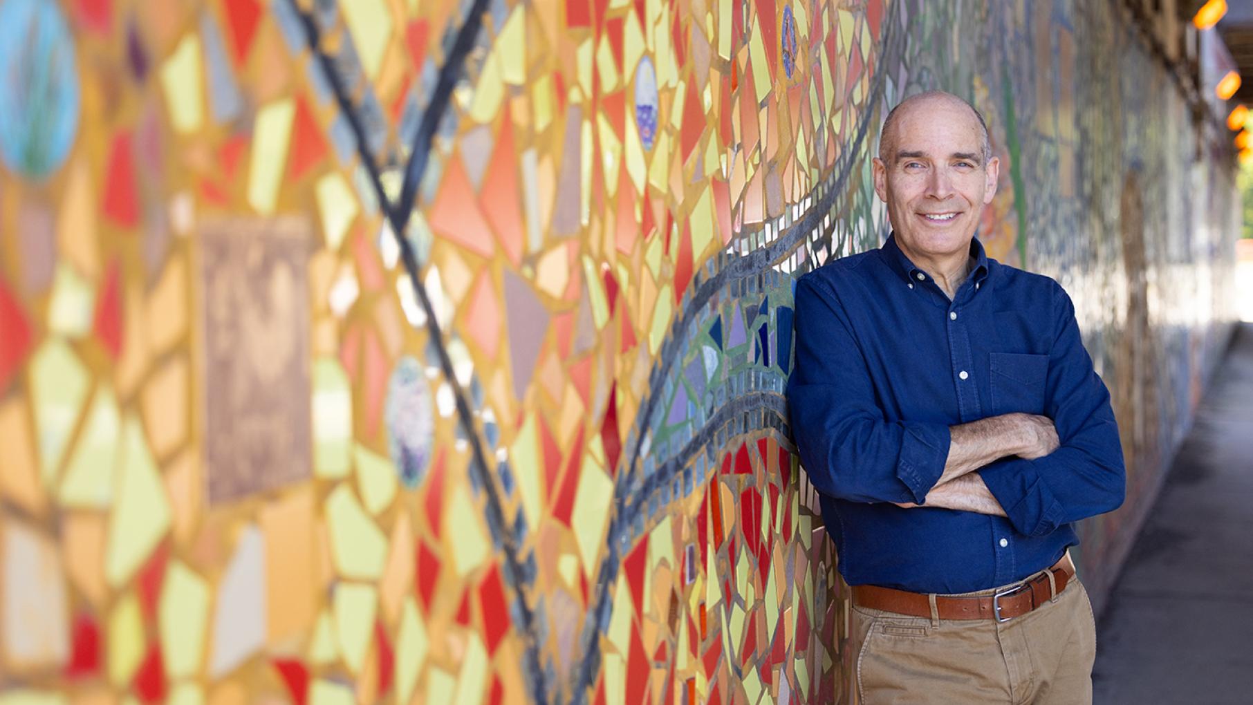 Geoffrey Baer stands in front of a colorful mosaic mural