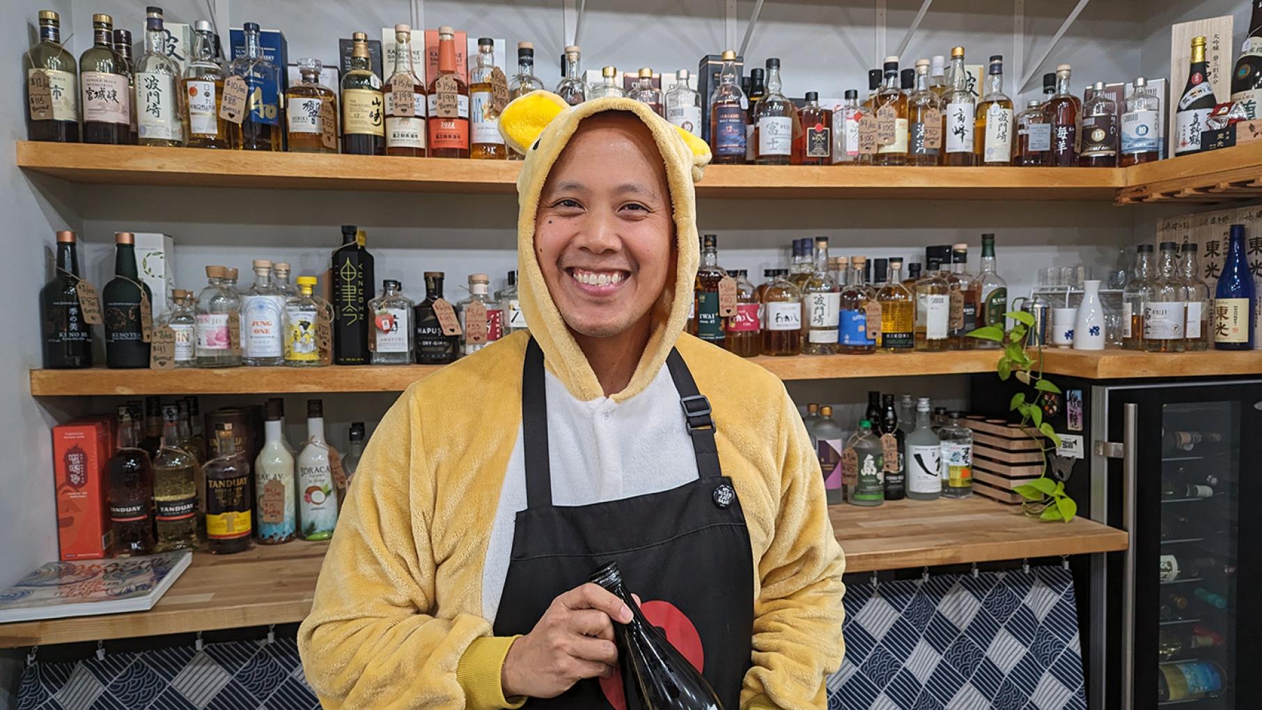 Jun-Jun Vichaikul poses in a onesie in front of shelves of liquor