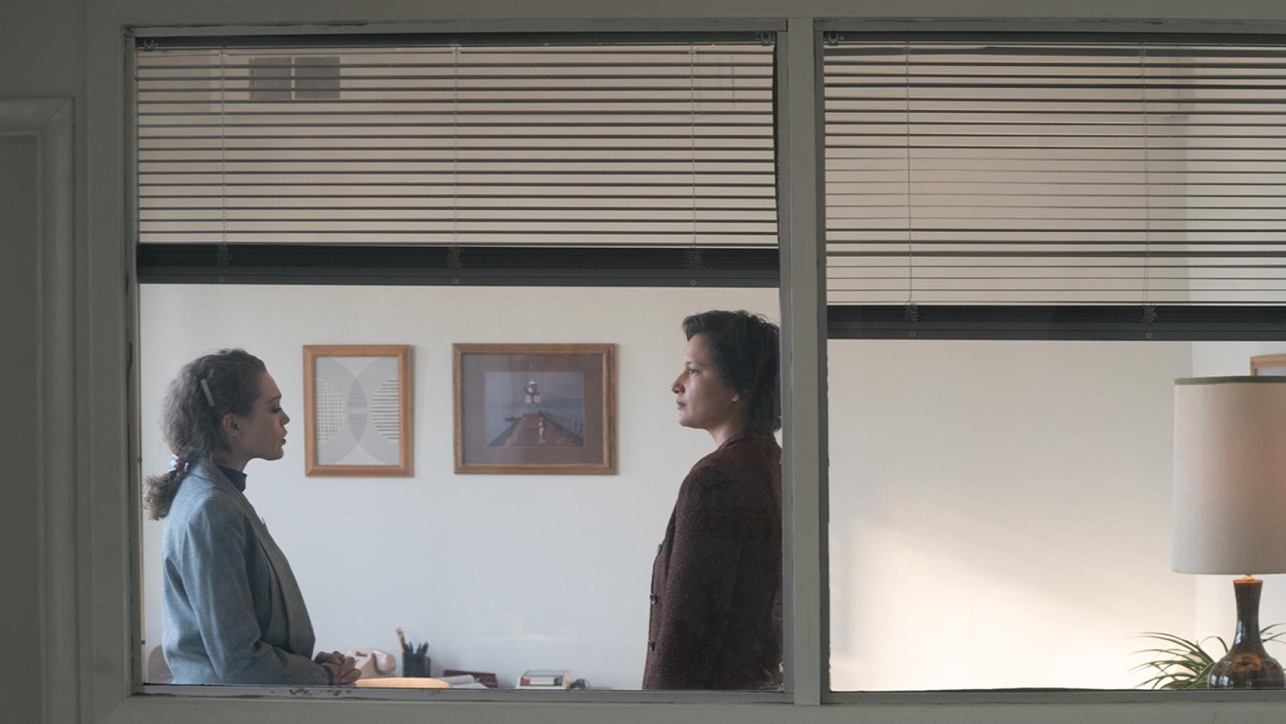 Bezhig and Adele seen standing facing each other in an office through a window