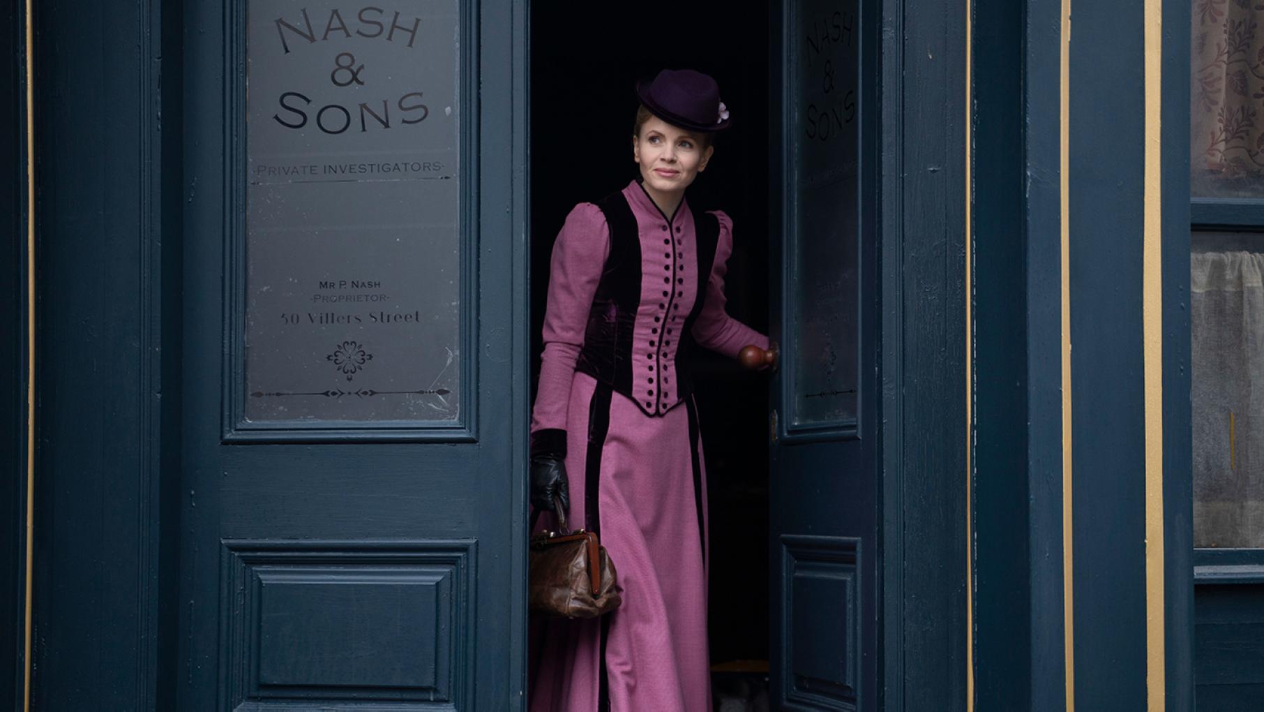 Miss Scarlet in a pink dress comes out of a door