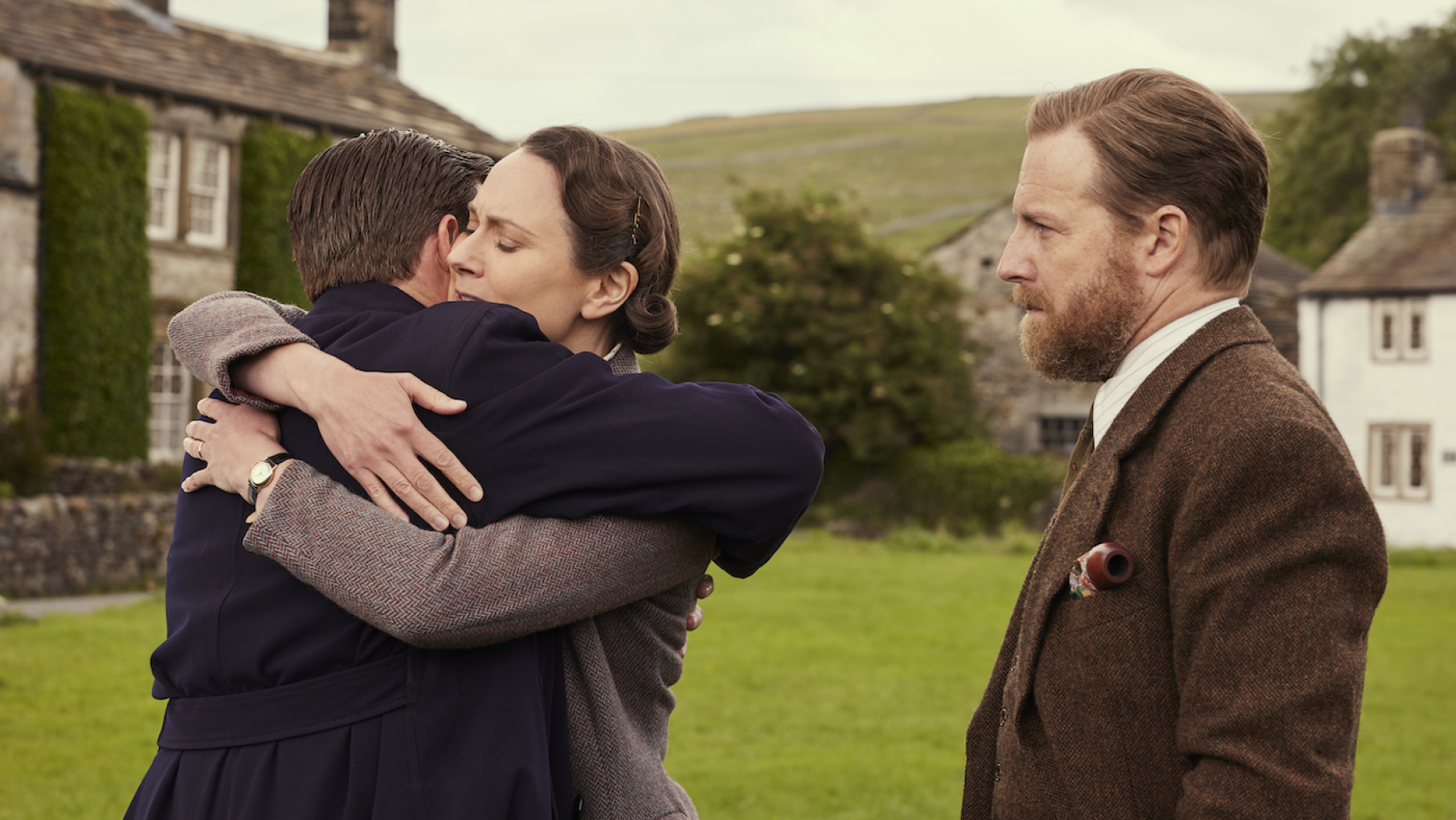 James hugs Mrs. Hall while Siegfried stands by in front of a grassy lawn and stone house