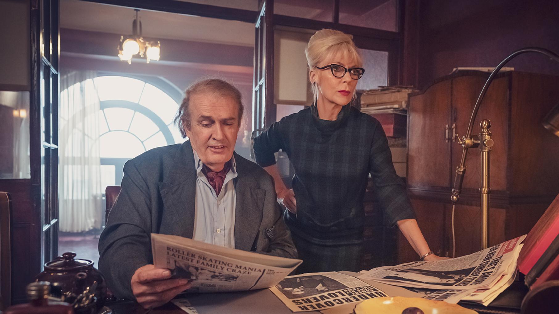 Brian tells Barbara to stay at home out of sight while she is suspended from her show. Credit: Potboiler Productions and Sky UK Limited Brian sits at a desk looking at a newspaper while Patsy stands next to him