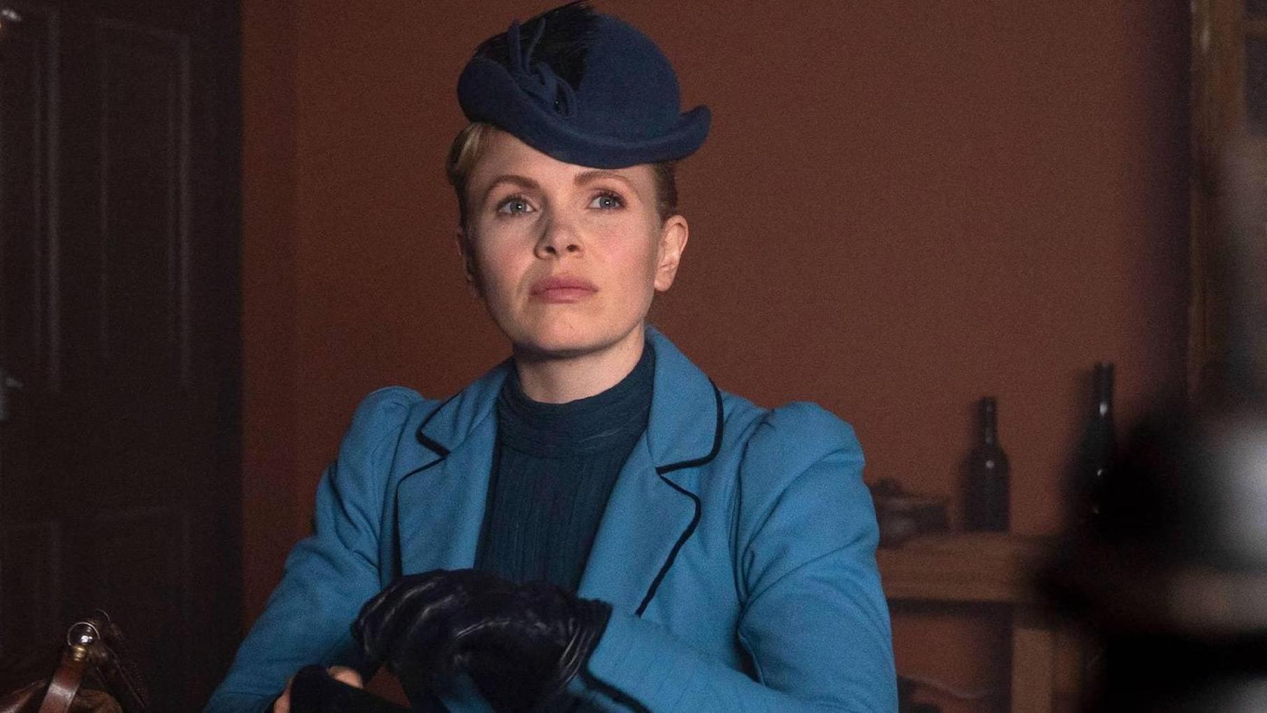 Eliza's business partner is wanted in connection with a murder. Credit: Masterpiece Eliza in a blue dress and hat sits and looks off camera