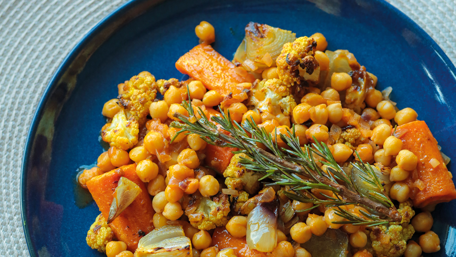 Diane Kochilas' Chickpea Stew with Honey-Roasted Cauliflower and Root Vegetables. Credit: Vasilis Stenos A blue plate of chickpea stew with vegetables and a sprig of rosemary