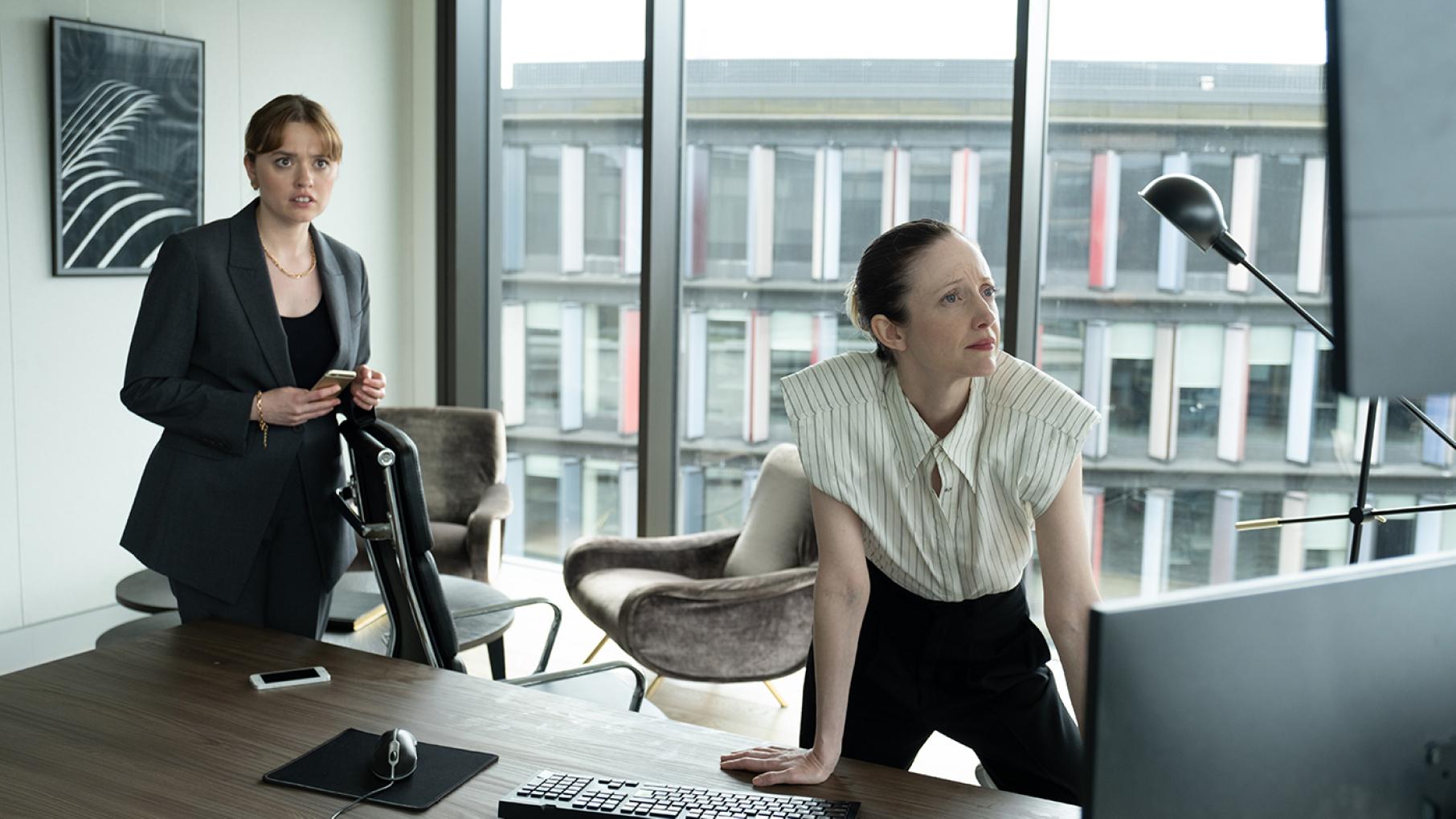 Maya and Alice look at computer screens on a desk in front of a wall of windows