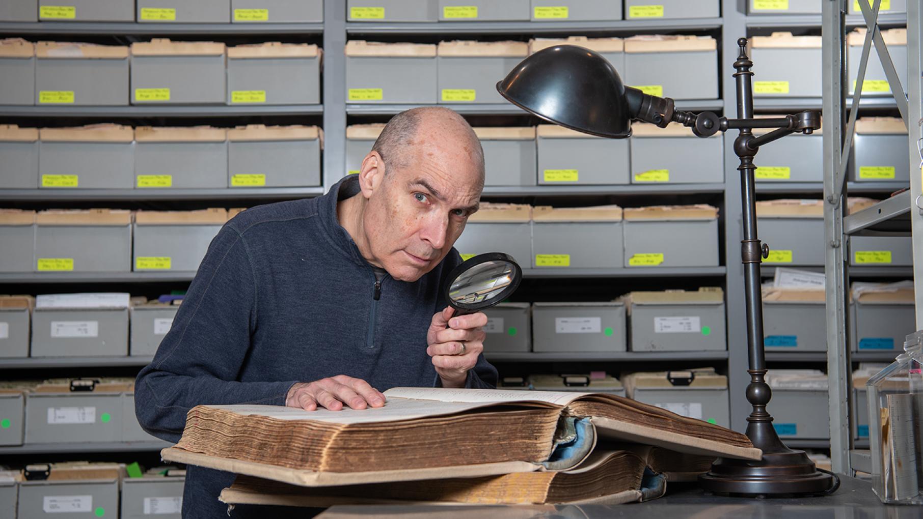 Geoffrey Baer investigates some of Chicago's most intriguing mysteries in a new special. Credit: Liz Farina Markel for WTTW Geoffrey Baer looks at an old record book under a lamp with a magnifying glass