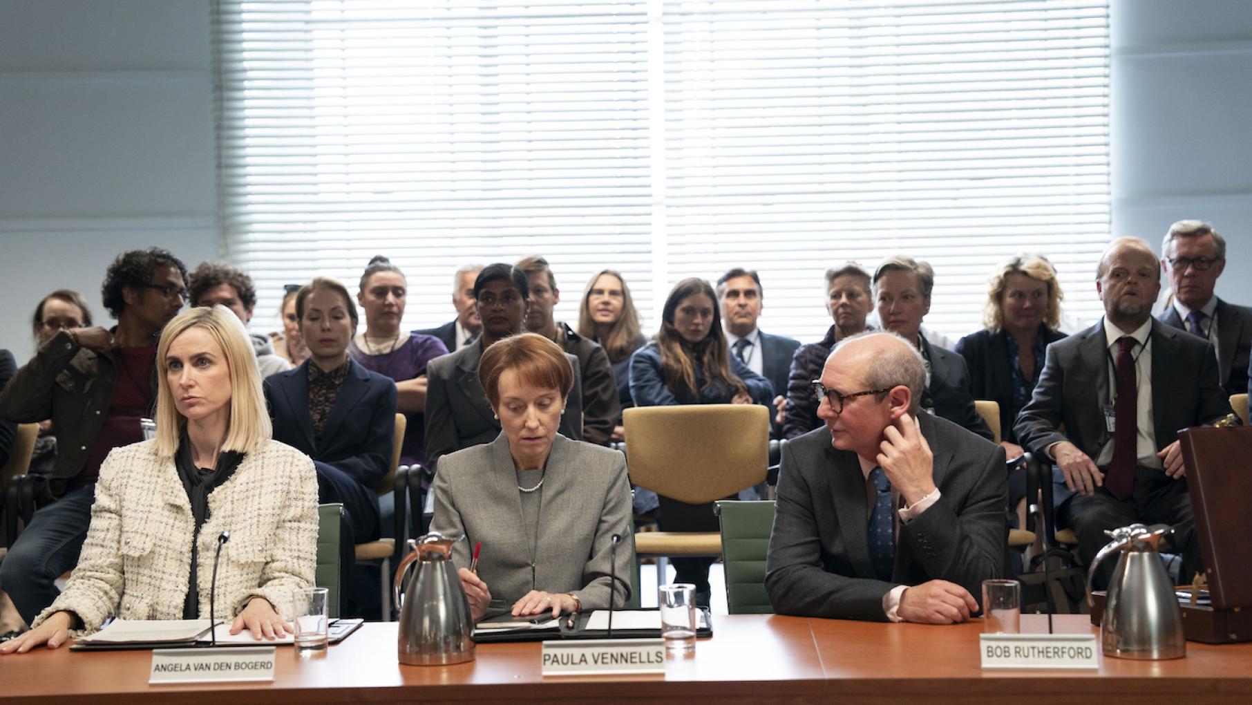 Angela, Paula, and Bob sit behind a table in front of a crowd at a hearing