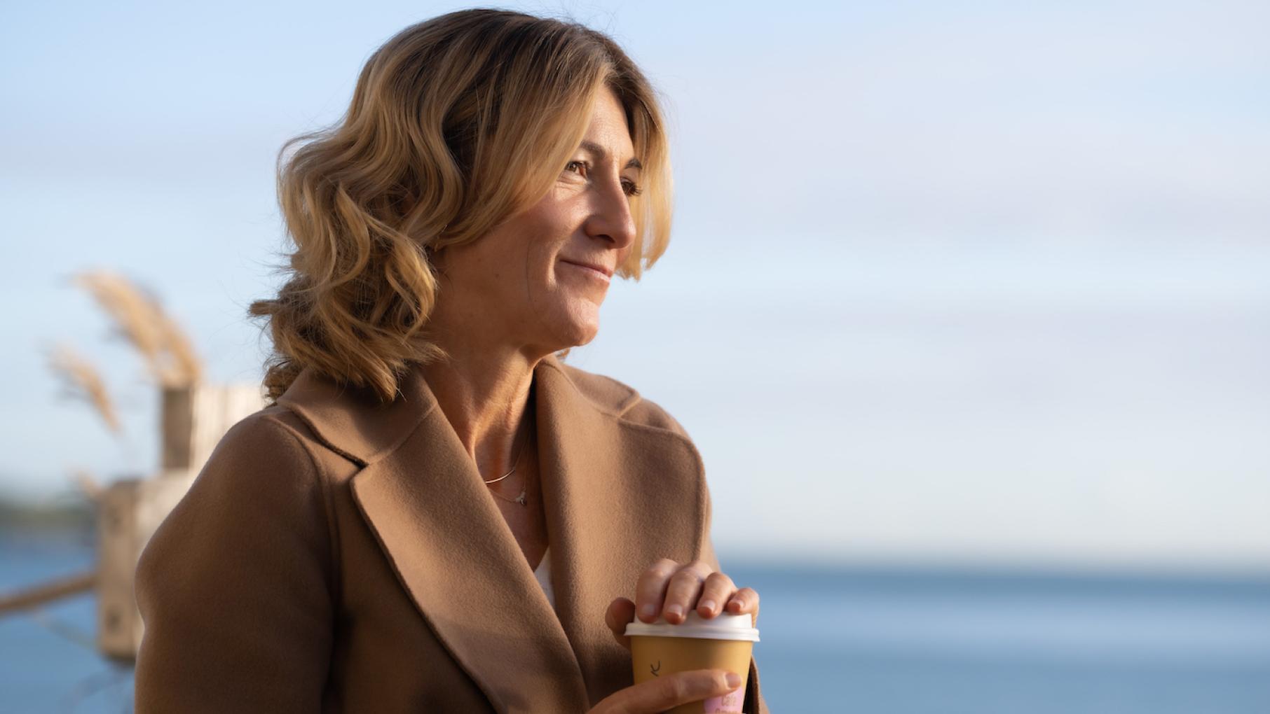 Rosaline is thrown together with her sister when their mother dies in an unexpected location. Credit: Monumental Television and Masterpiece Rosaline holds a cup of coffee and smiles in front of the ocean