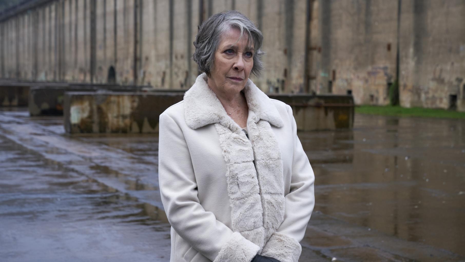 Maggie Lynch stands in a rainy industrial area in a coat