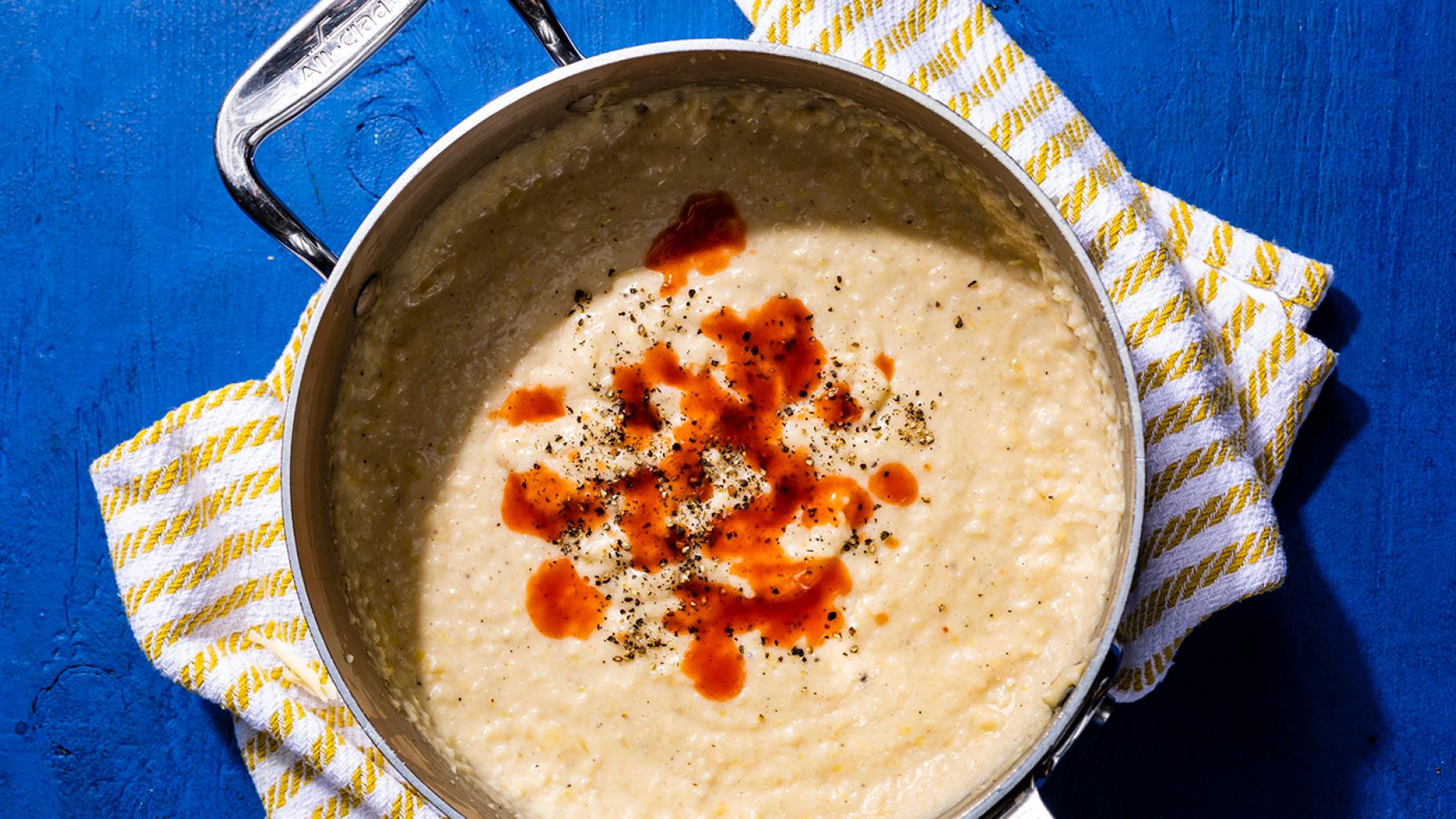 Credit: Steve Klise for Cook's Country A pot of cheesy grits topped with hot sauce on a blue background