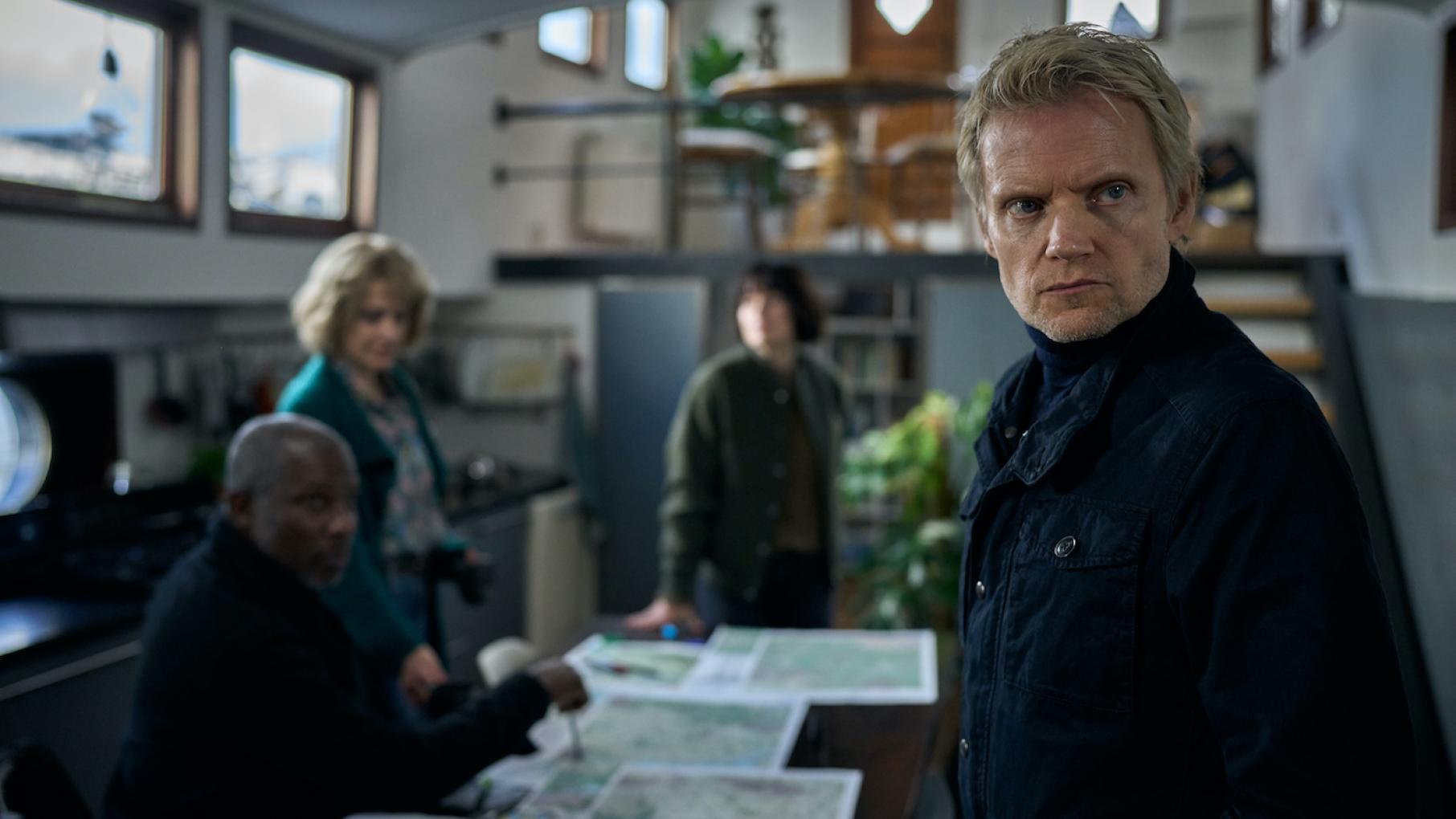 Van der Valk in the foreground while others sit and stand behind him in a houseboat