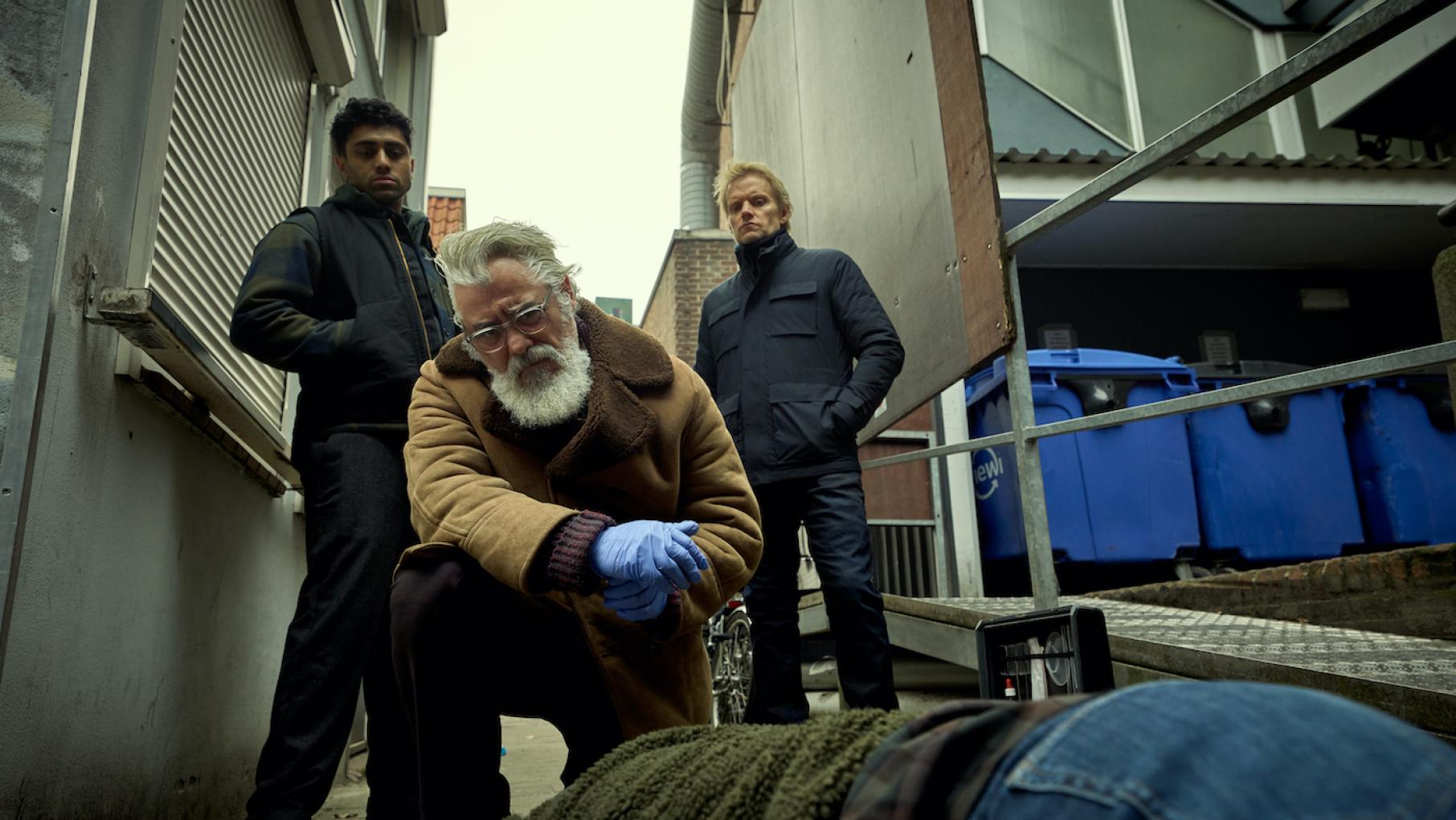 The team investigates the seemingly professional killing of two scientists researching cancer. Credit: All3Media International and Masterpiece Eddie, Hendrik, and Van der Valk look at a body on the ground outside