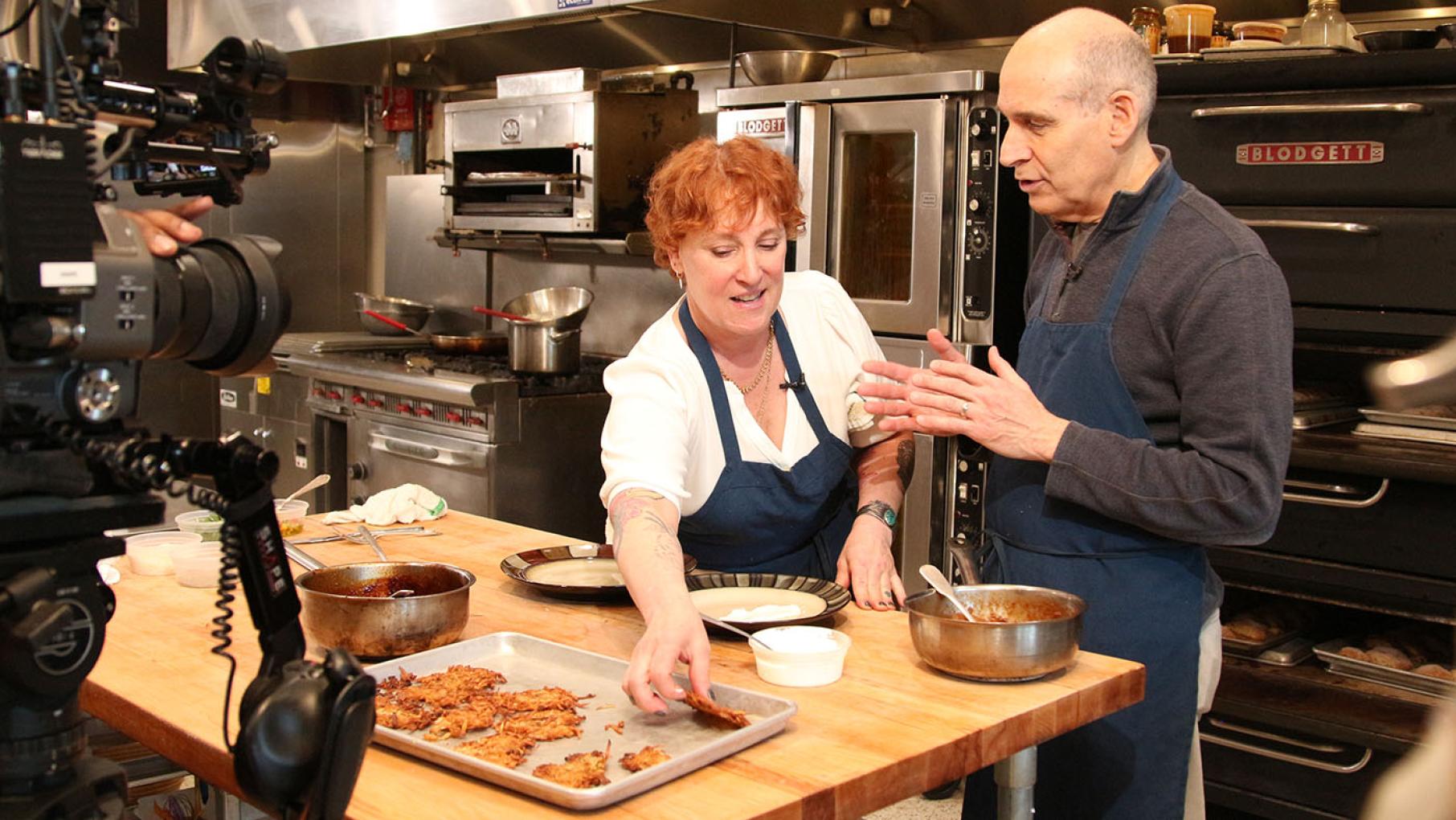 Geoffrey Baer and Mindy Segal in a restaurant kitchen making latkes with a camera in the frame