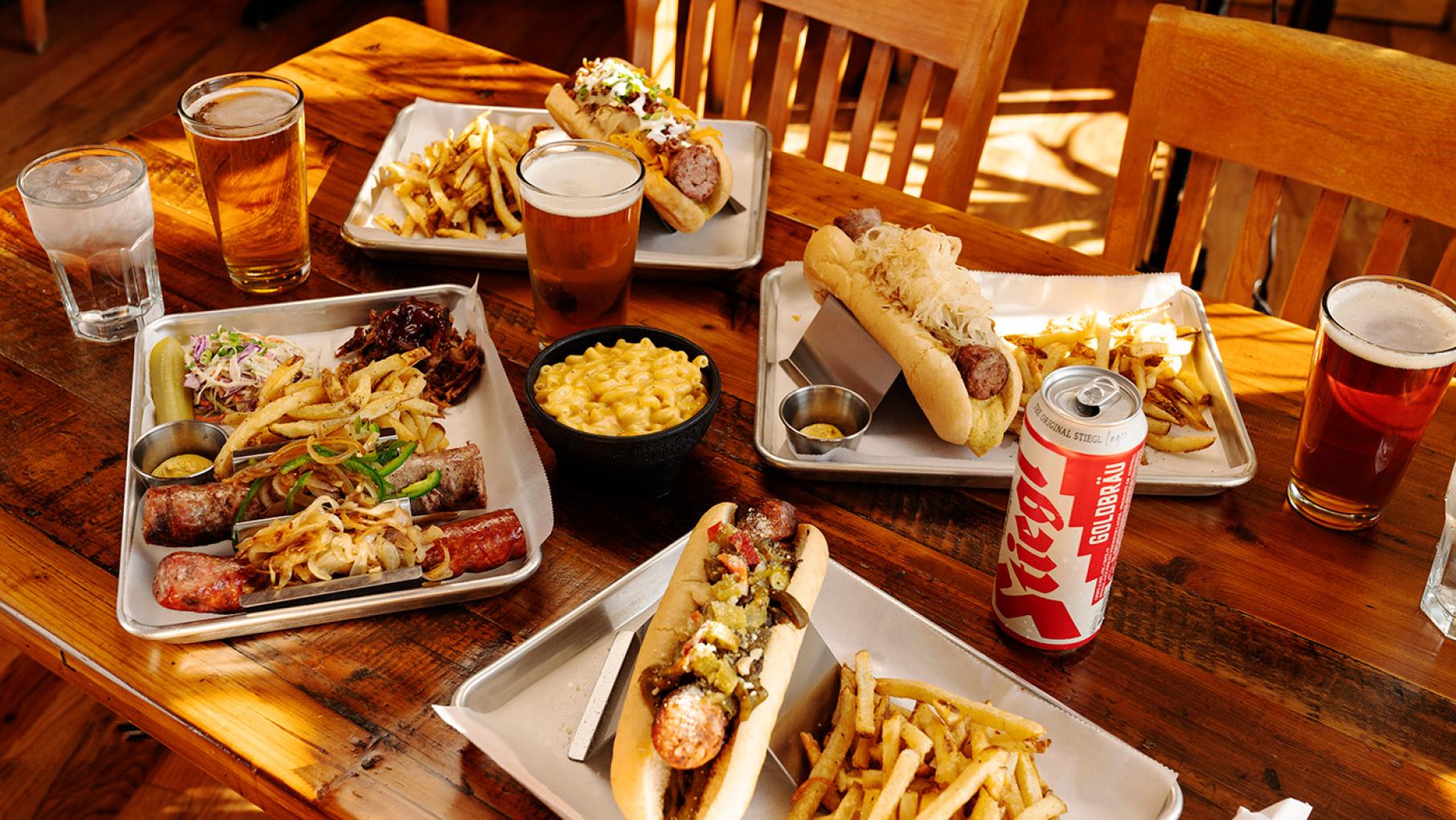 A wooden table with trays of sausage, fries, mac and cheese, and beers in cans and glasses