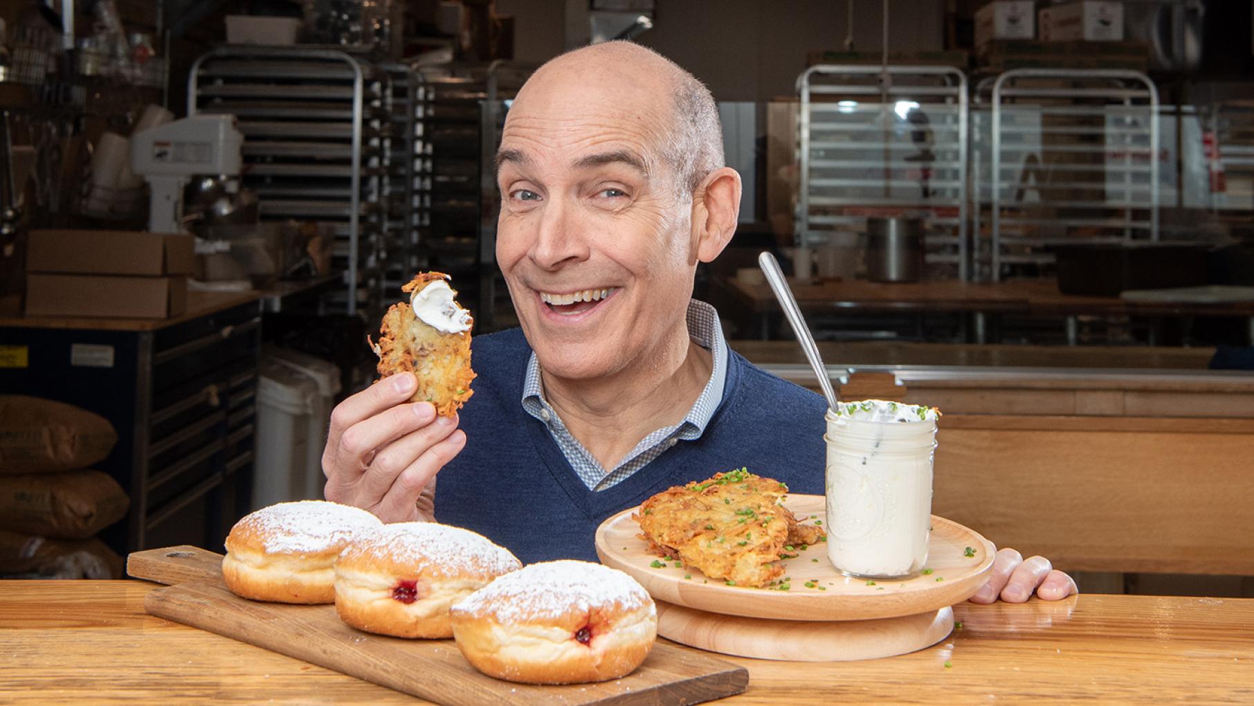 In 'A Celebration of Hanukkah,' Geoffrey Baer explores the joyous holiday and its traditions. Credit: Liz Farina Markel for WTTW Geoffrey Baer smiles behind a tray of sufganiyot and latkes and holds up a latke