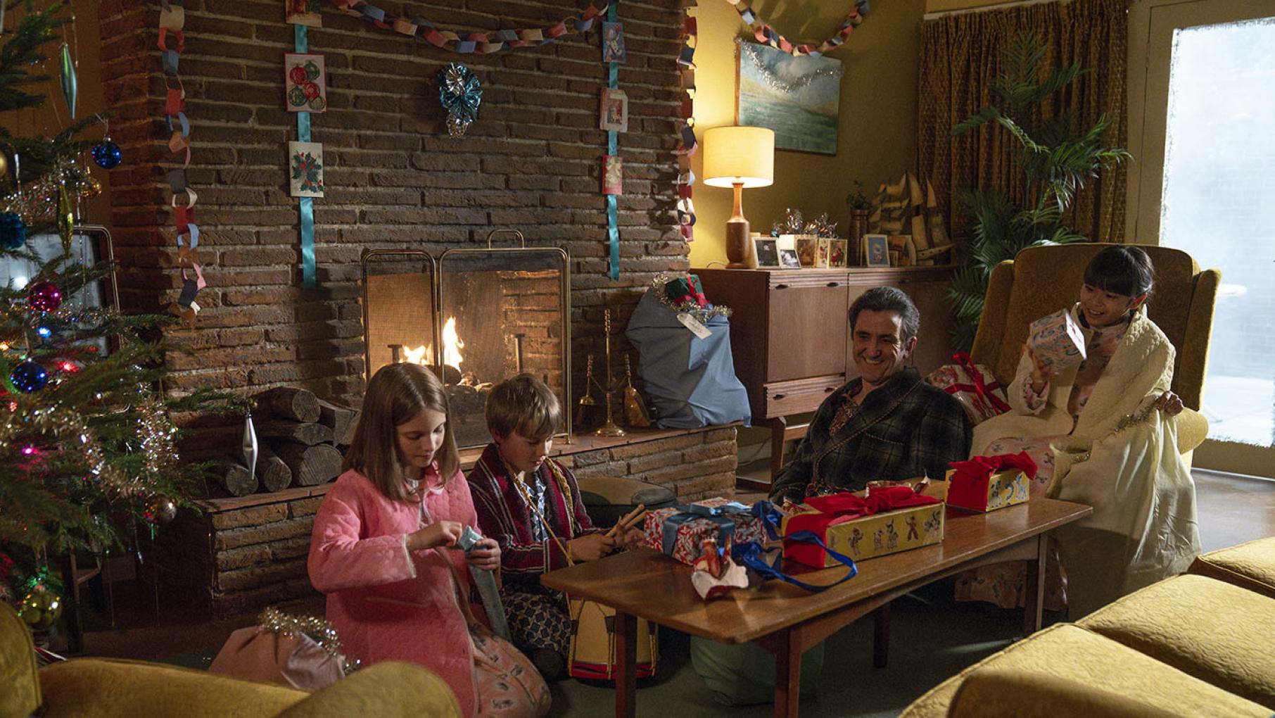 Angela Turner (Alice Brown), Teddy Turner (Edward Shaw), Dr. Patrick Turner (Stephen McGann), and May Tang (April Rae Hoang) in the Call the Midwife 2024 holiday special. Credit: BBC Studios Distribution Limited Three children and a man sit in a living room opening Christmas presents