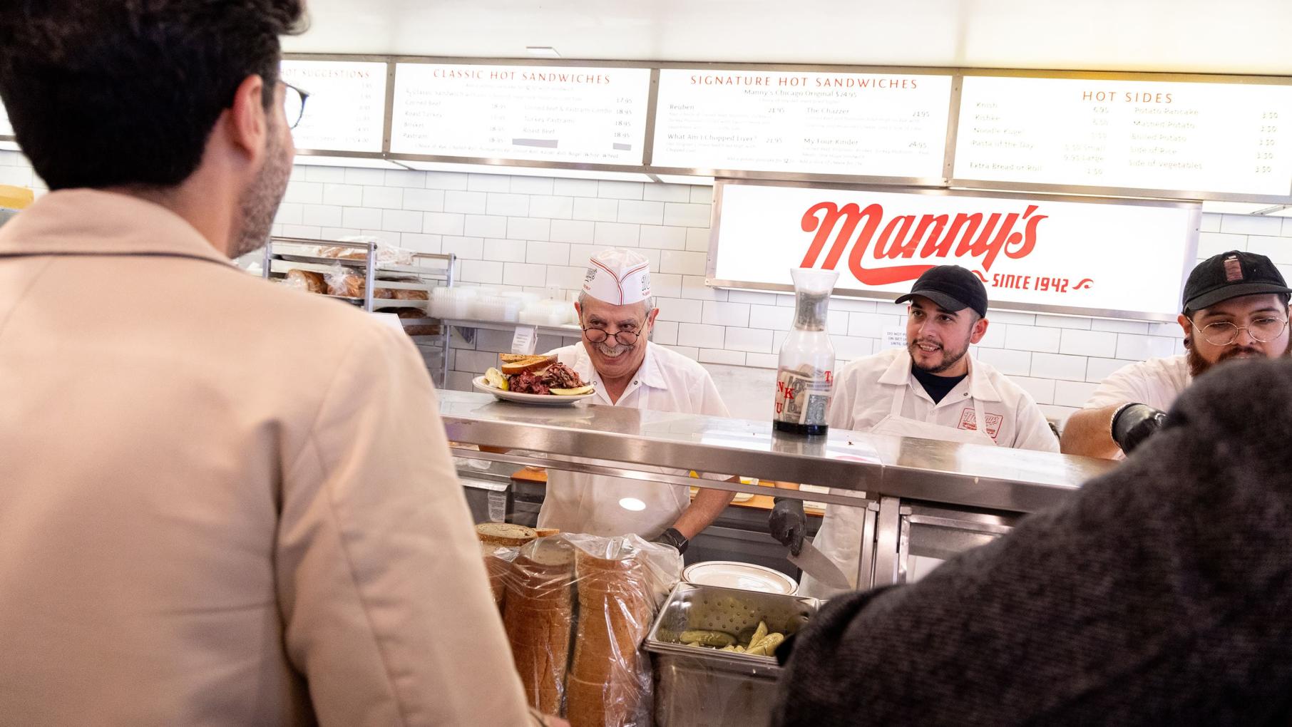 A customer ordering food at a counter with servers behind, featuring a sign for "Manny's."